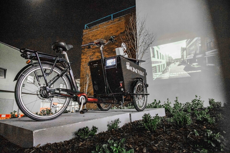 A black cargo bike is displayed on a platform outdoors at night. Behind the bike, a black-and-white video is projected onto a wall, and nearby plants add greenery to the scene.